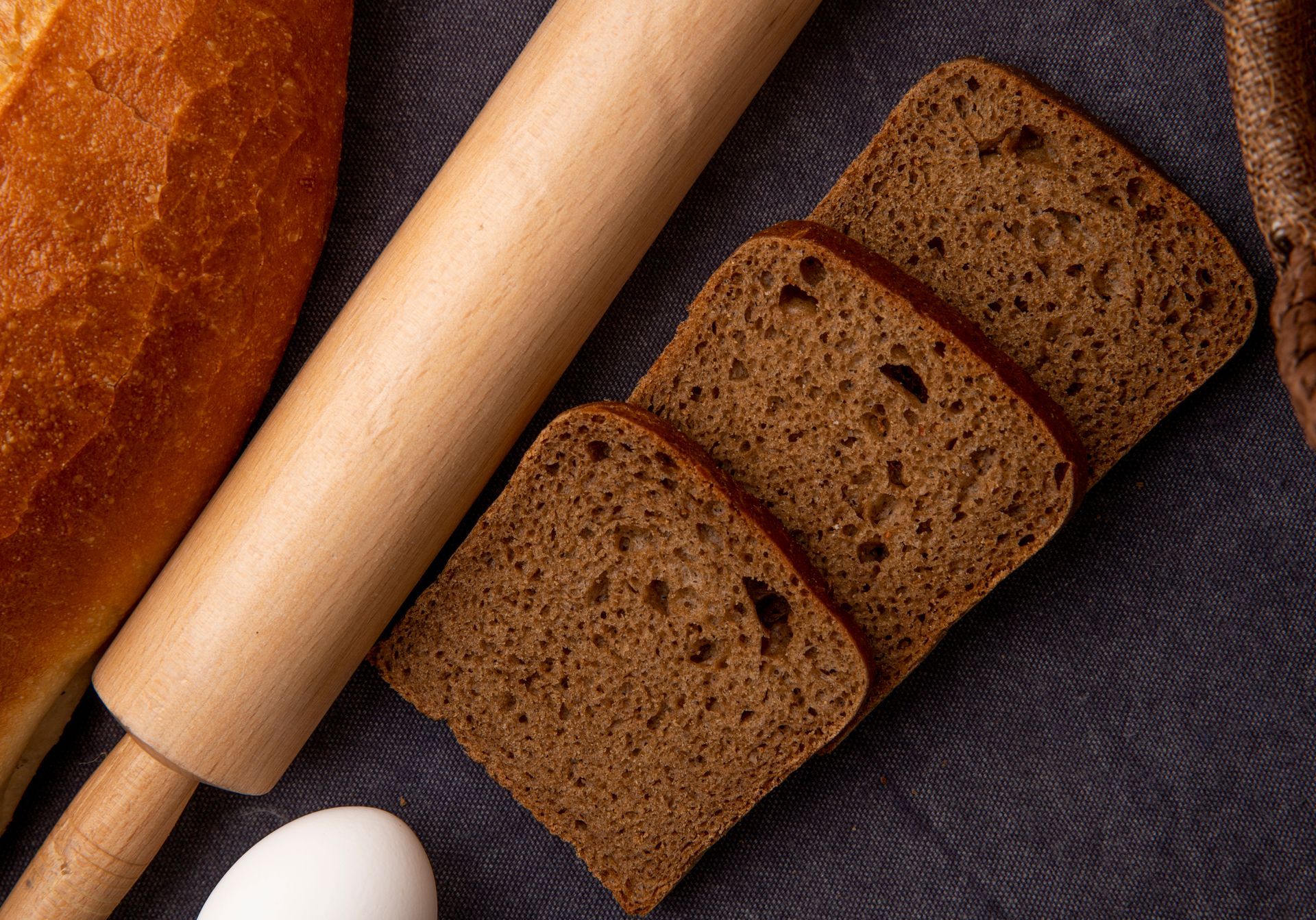Sliced brown bread, rolling pin, bread loaf, and egg on a dark cloth surface.