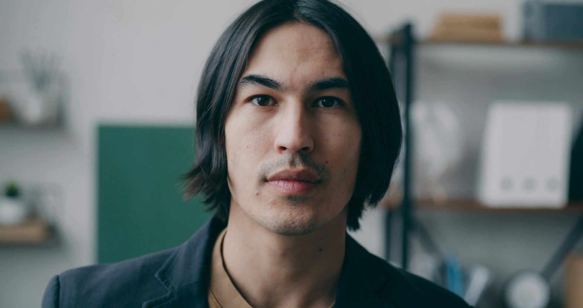 Man with long, dark hair and a neutral expression, wearing a dark blazer, in an office setting.