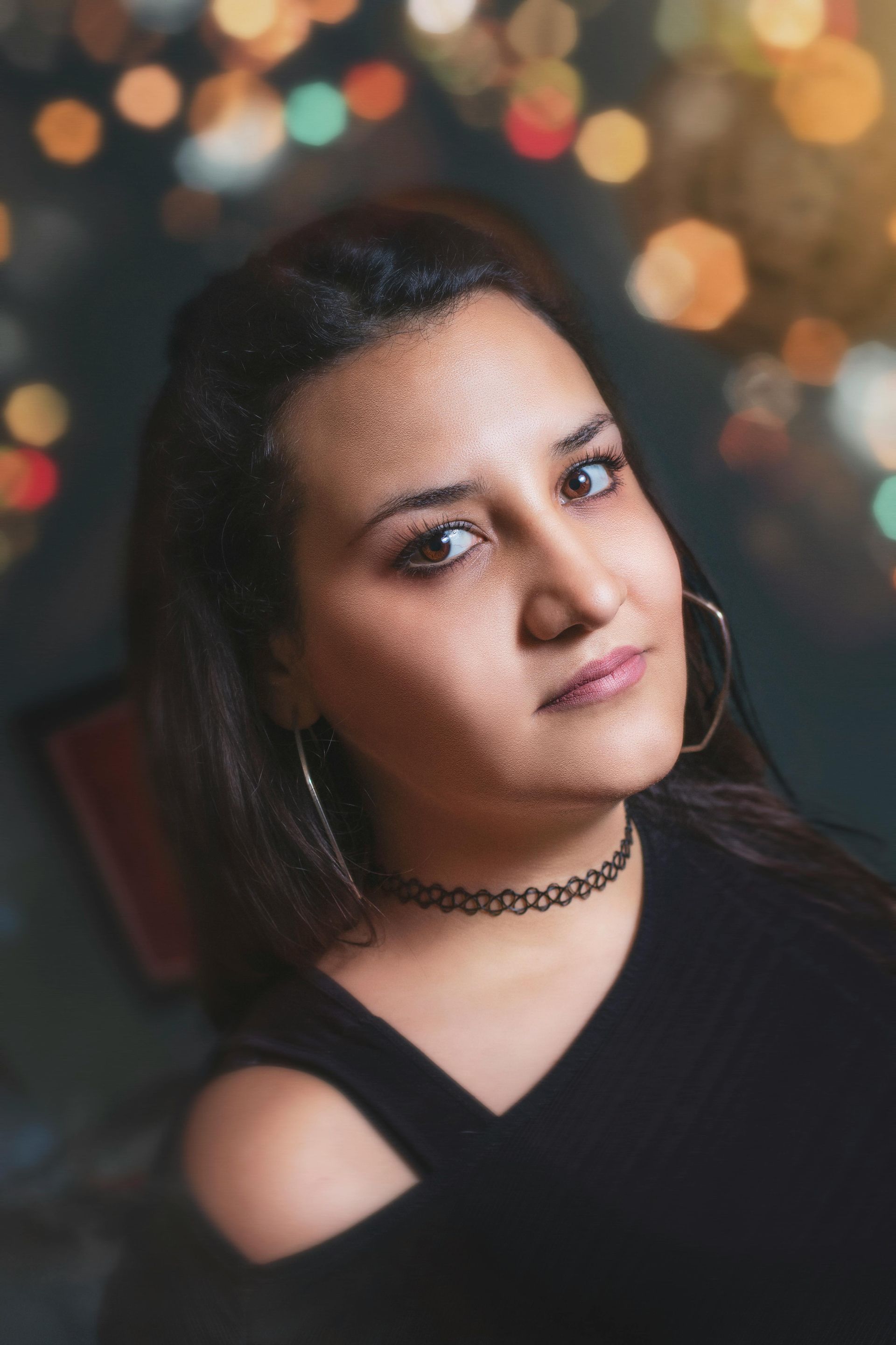 Woman with dark hair, looking at the camera, wearing a black top and choker necklace, with bokeh background.