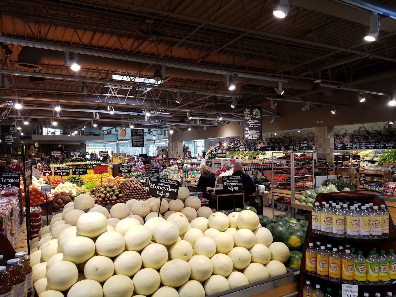 Grocery store interior with produce display of melons; shoppers browse.