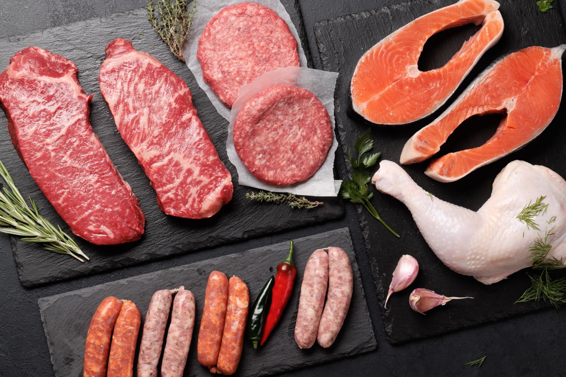 Assortment of raw meats on black slate: steaks, burgers, salmon fillets, chicken leg, sausages, and spices.