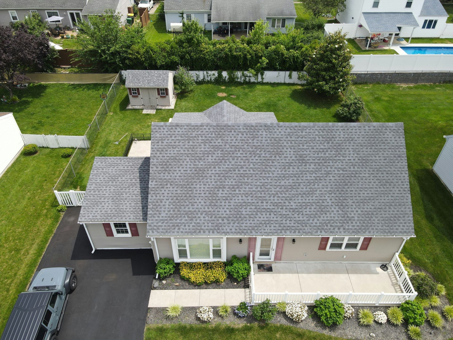 An aerial view of a gray, single-story suburban house with a dark roof, front porch, and a driveway with a parked car.