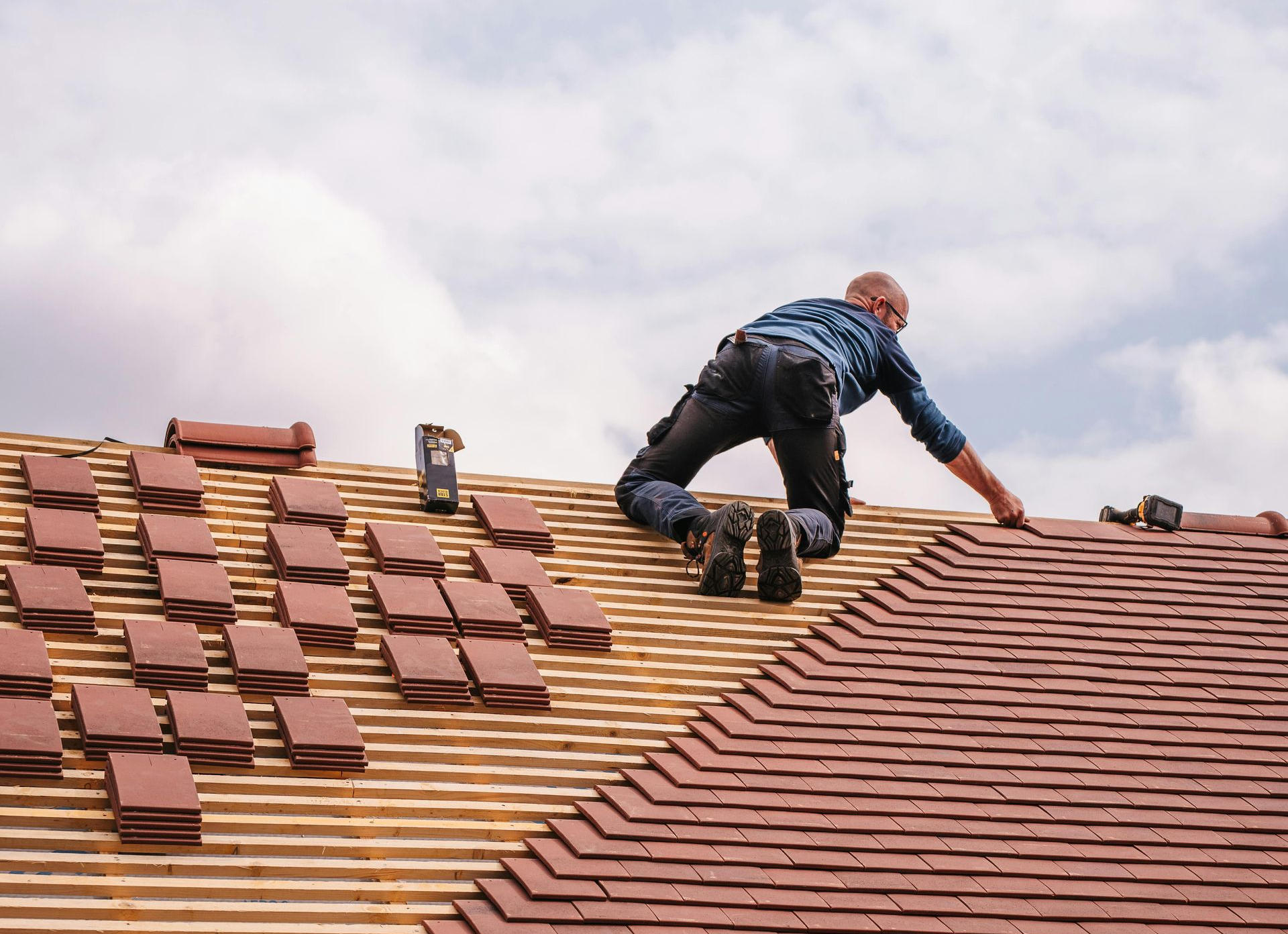 A roofer works on a sloped roof, installing clay tiles onto wooden battens against a cloudy sky.