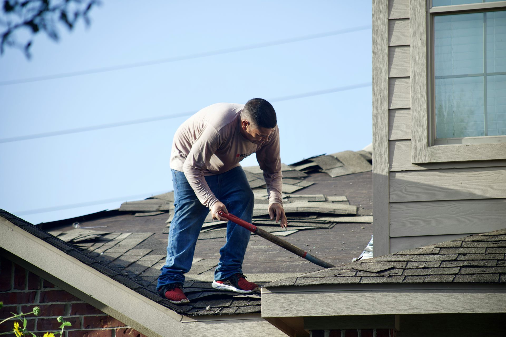 A person wearing a long-sleeved shirt and jeans uses a tool to remove old shingles from a house roof.