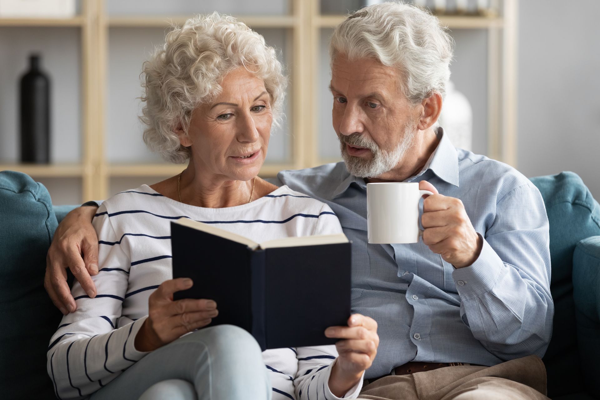 An elderly couple is sitting on a couch reading a book and drinking coffee.