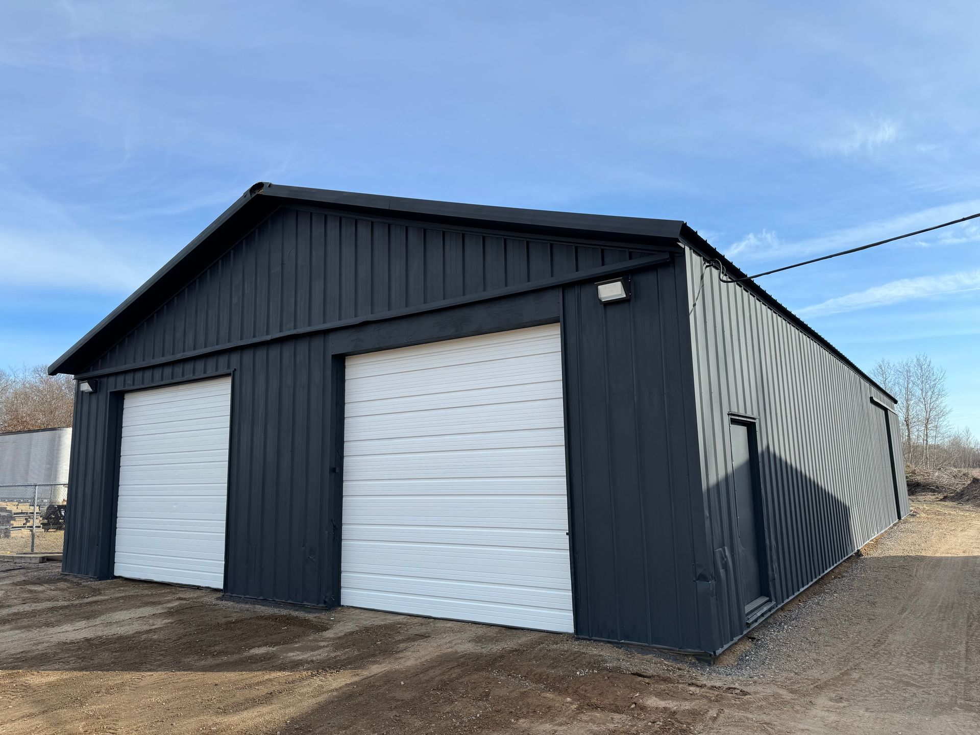 A black garage with two white garage doors on a dirt road.