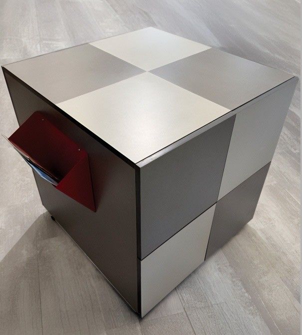 Objeto plateado con forma de cubo, con la parte superior a cuadros y una ranura roja en el lateral, sobre un suelo gris.