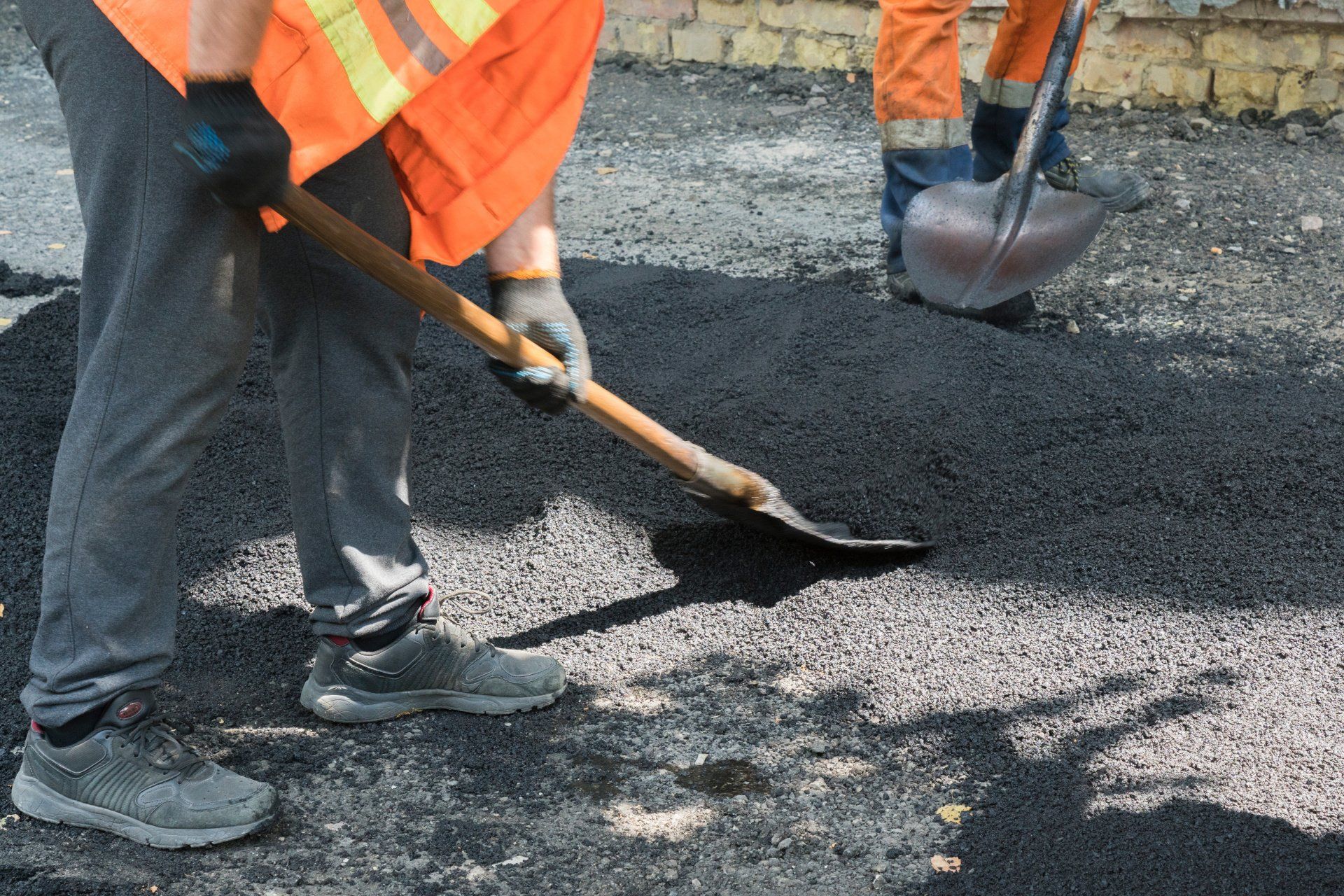 A worker in a yellow vest is laying asphalt to repair a road under the bright rays of the sun.