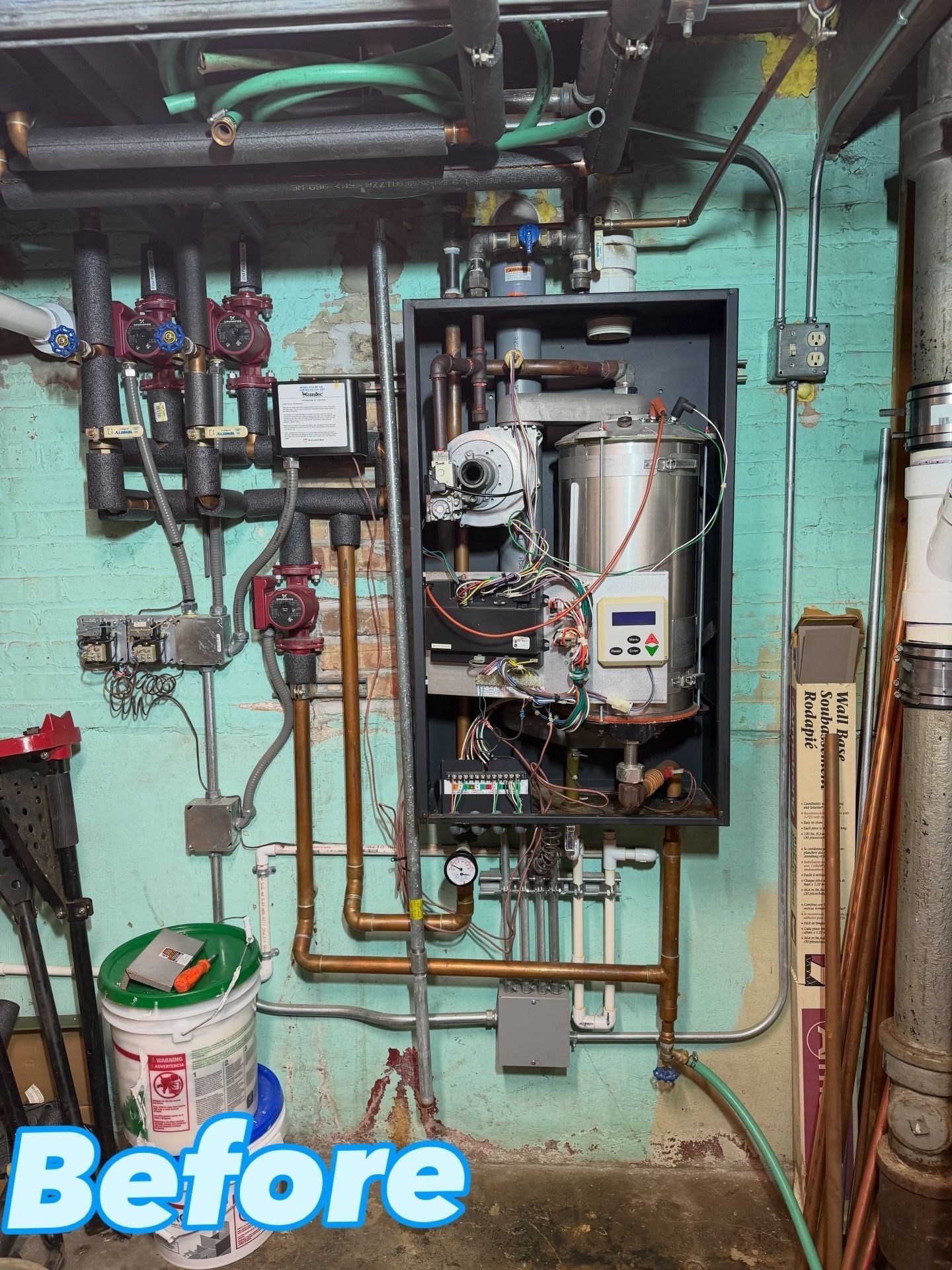 A before photo of a boiler and related piping installed on a painted concrete wall.