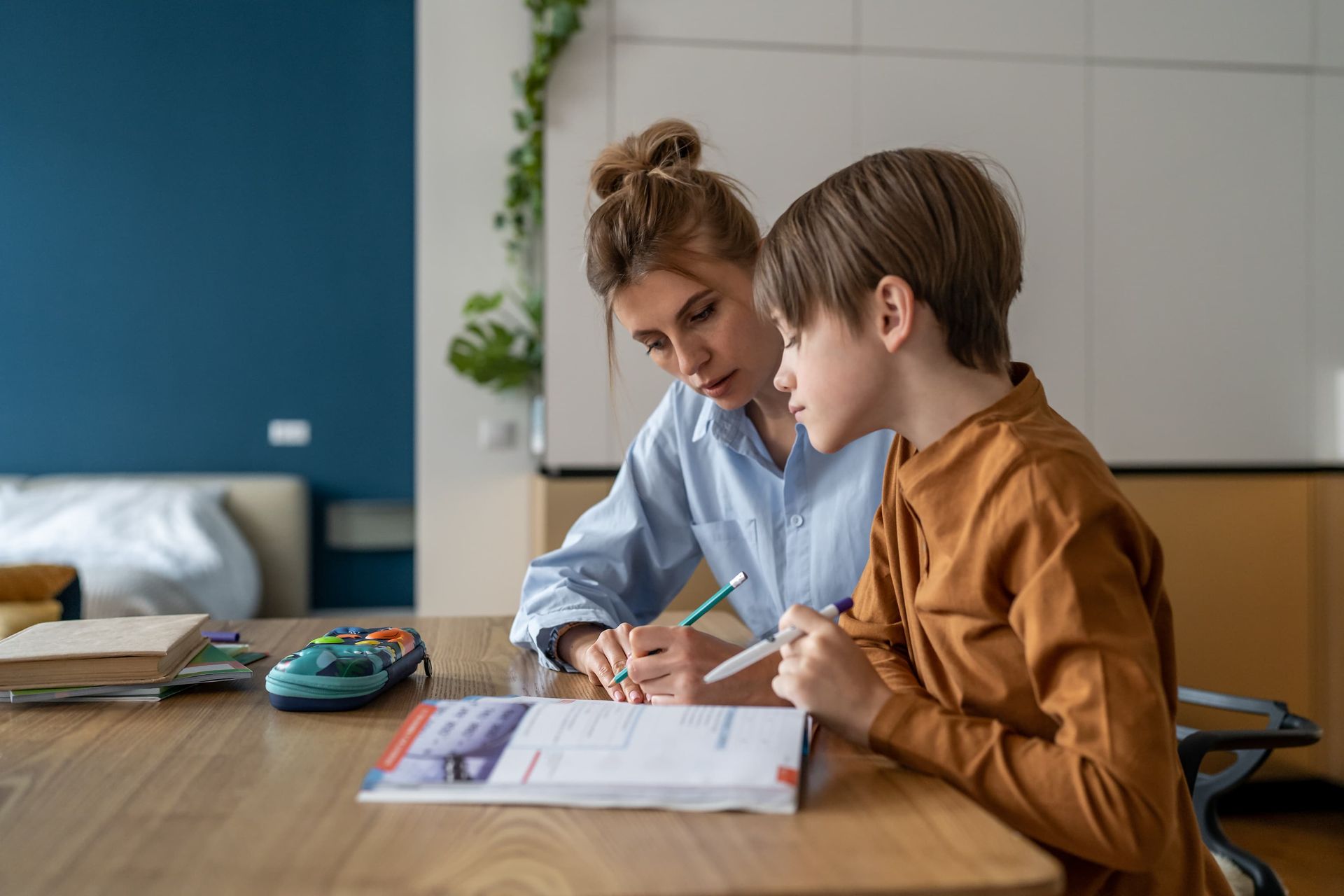 Woman assisting a child with homework at a wooden table. They are both writing in a book.
