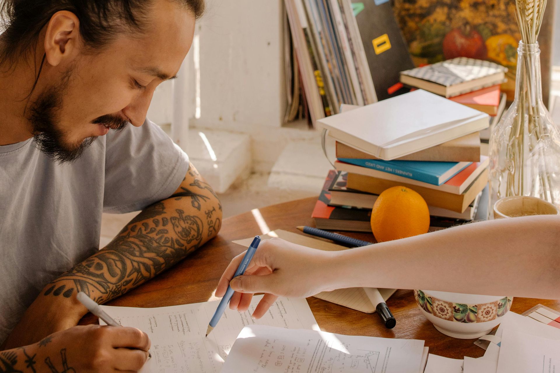 Two people work together at a wooden table, writing on papers amidst a stack of books and an orange.