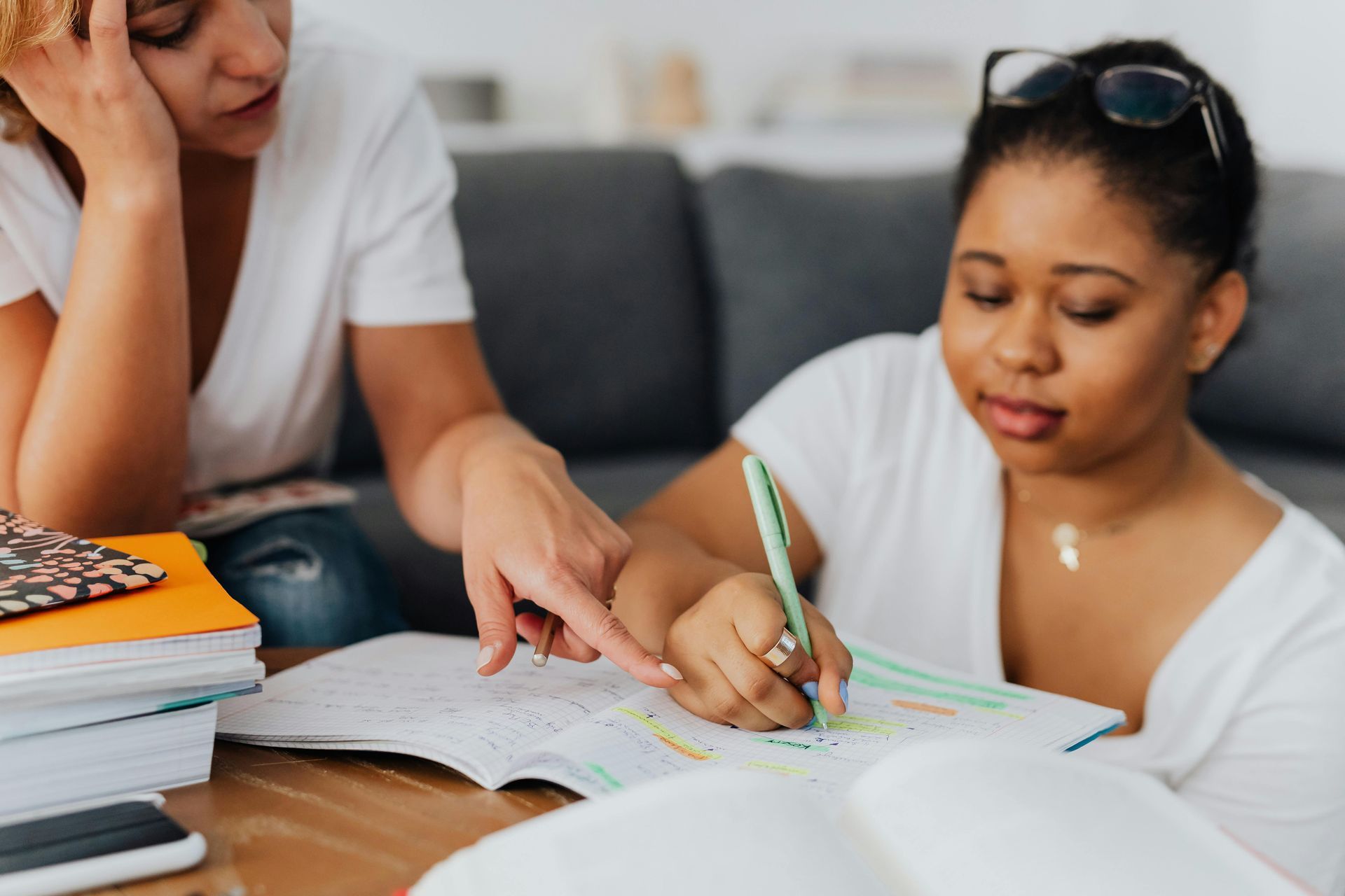 Two people study together at a table, one pointing to notes in an open book while the other writes with a green pen.