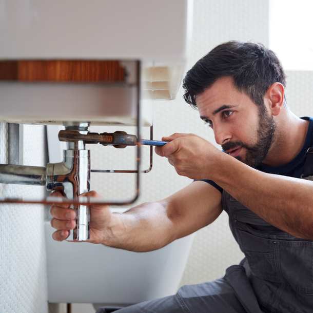 Plumber with a wrench fixing pipes under a white sink in a bathroom.