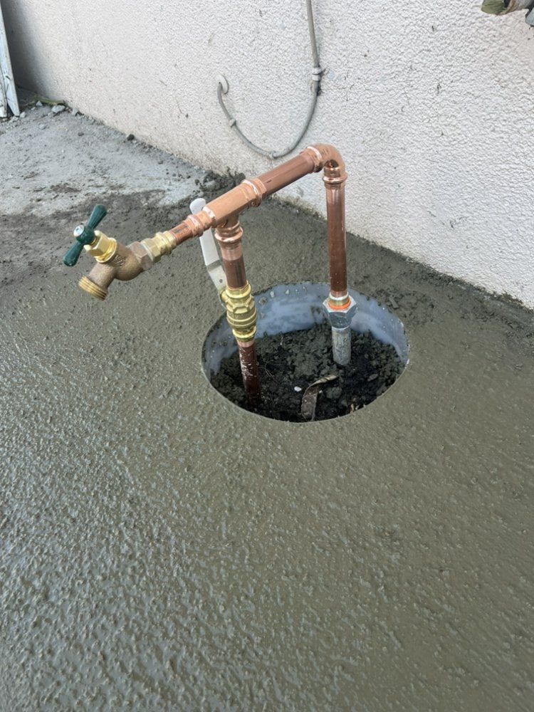 Copper pipes and faucet rising from a hole in wet concrete, near a white wall.