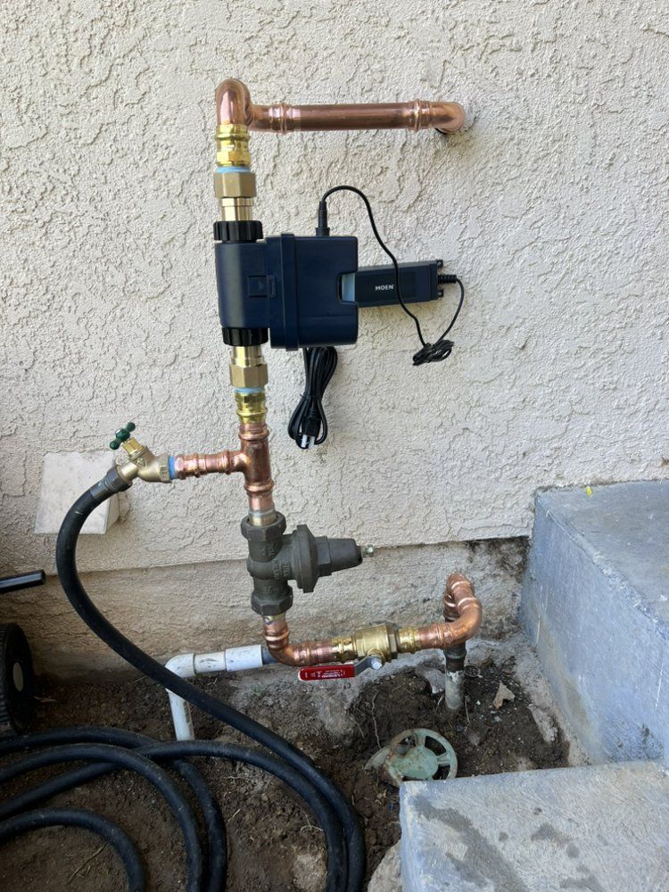 Copper pipes and fixtures for irrigation system attached to wall. Includes a pressure regulator and a black electronic device.