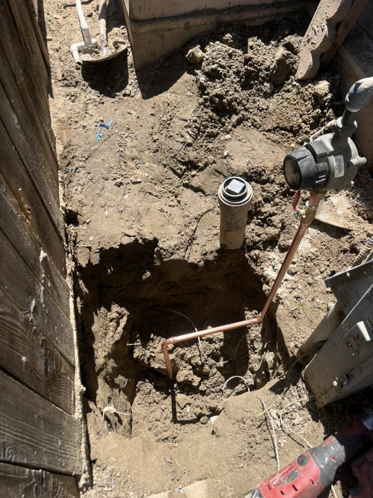A trench in the dirt with copper piping, water meter, and sprinkler head, next to a wooden fence.