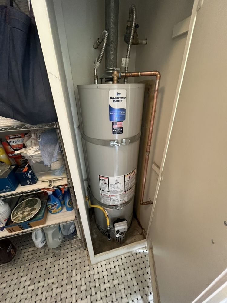 A tall water heater in a white closet. Copper and gray pipes connect to the tank.