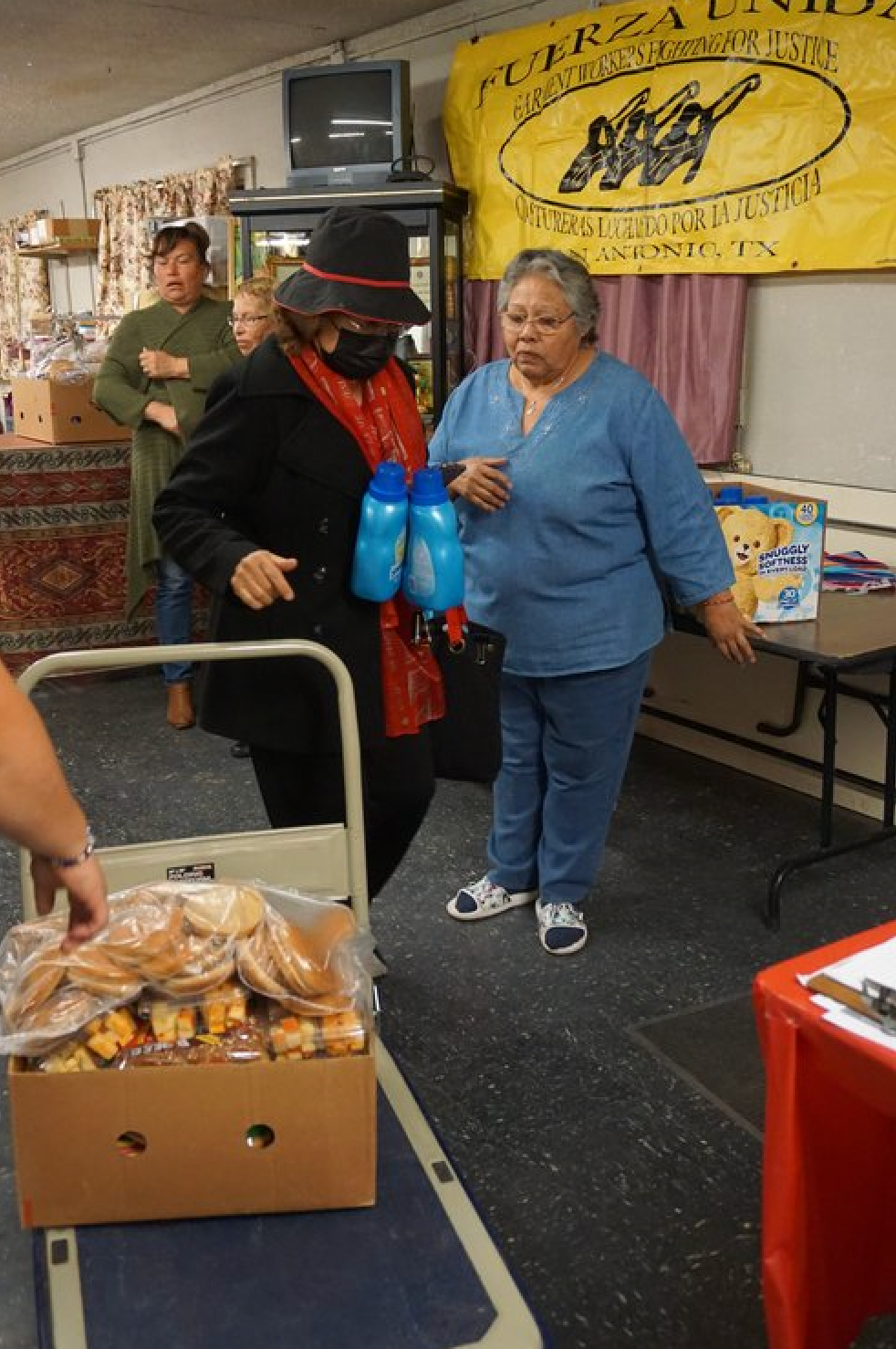 Two women are standing in a room with a box of bread on a cart.