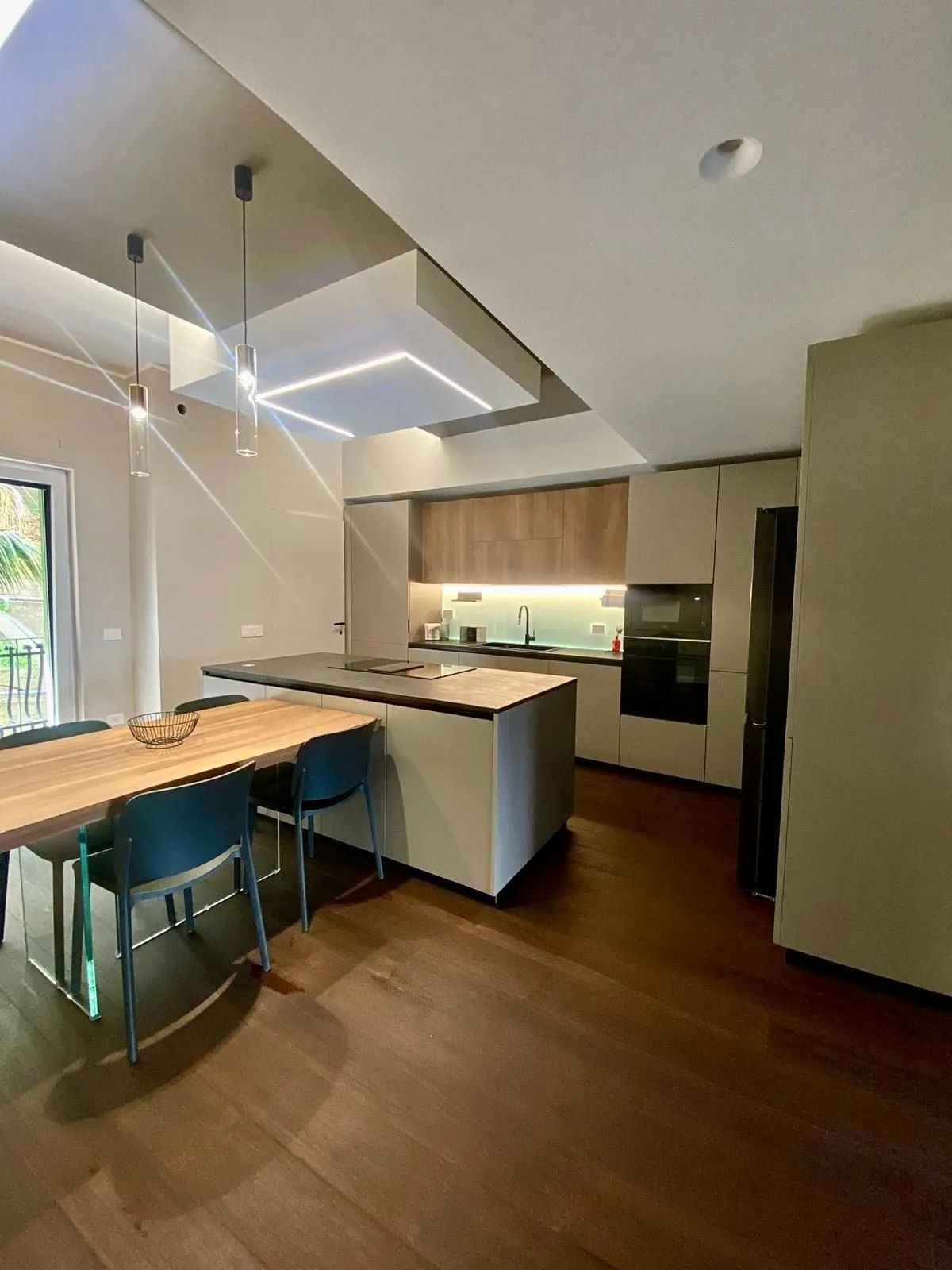 Cucina moderna con pavimento in legno, isola con posti a sedere, lampade a sospensione e forno a incasso.