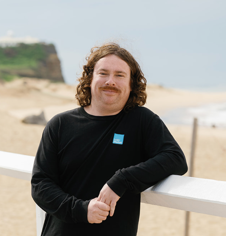 Man with curly hair and mustache leans on a railing at the beach, wearing a black shirt.