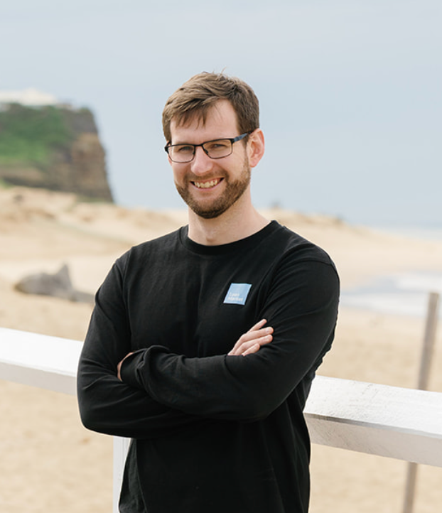 Man with glasses smiles, arms crossed, black shirt, beach background.