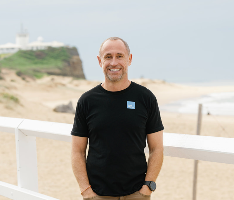 Man in black t-shirt smiles at camera, standing on a beach with a white fence. A lighthouse is in the background.