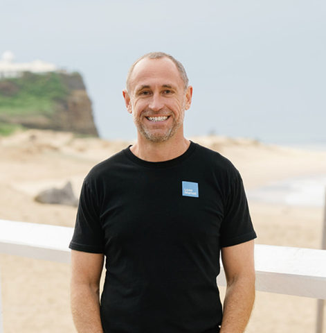 Man in black t-shirt smiles at the camera on a beach.