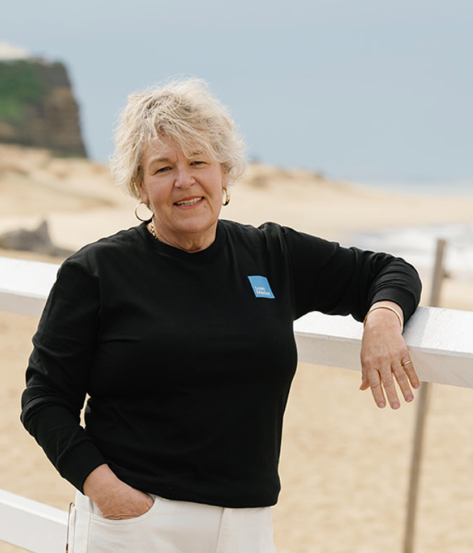 Woman in black shirt leaning on a white fence at the beach, smiling, hand in pocket.