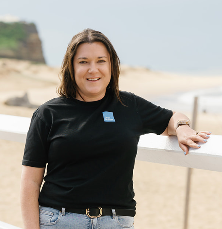 Woman in black t-shirt leaning on a white railing at the beach, smiling.