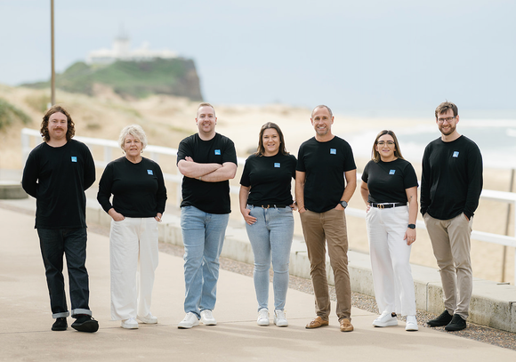 Group of seven people, all wearing black shirts with a logo, standing on a path near a beach.