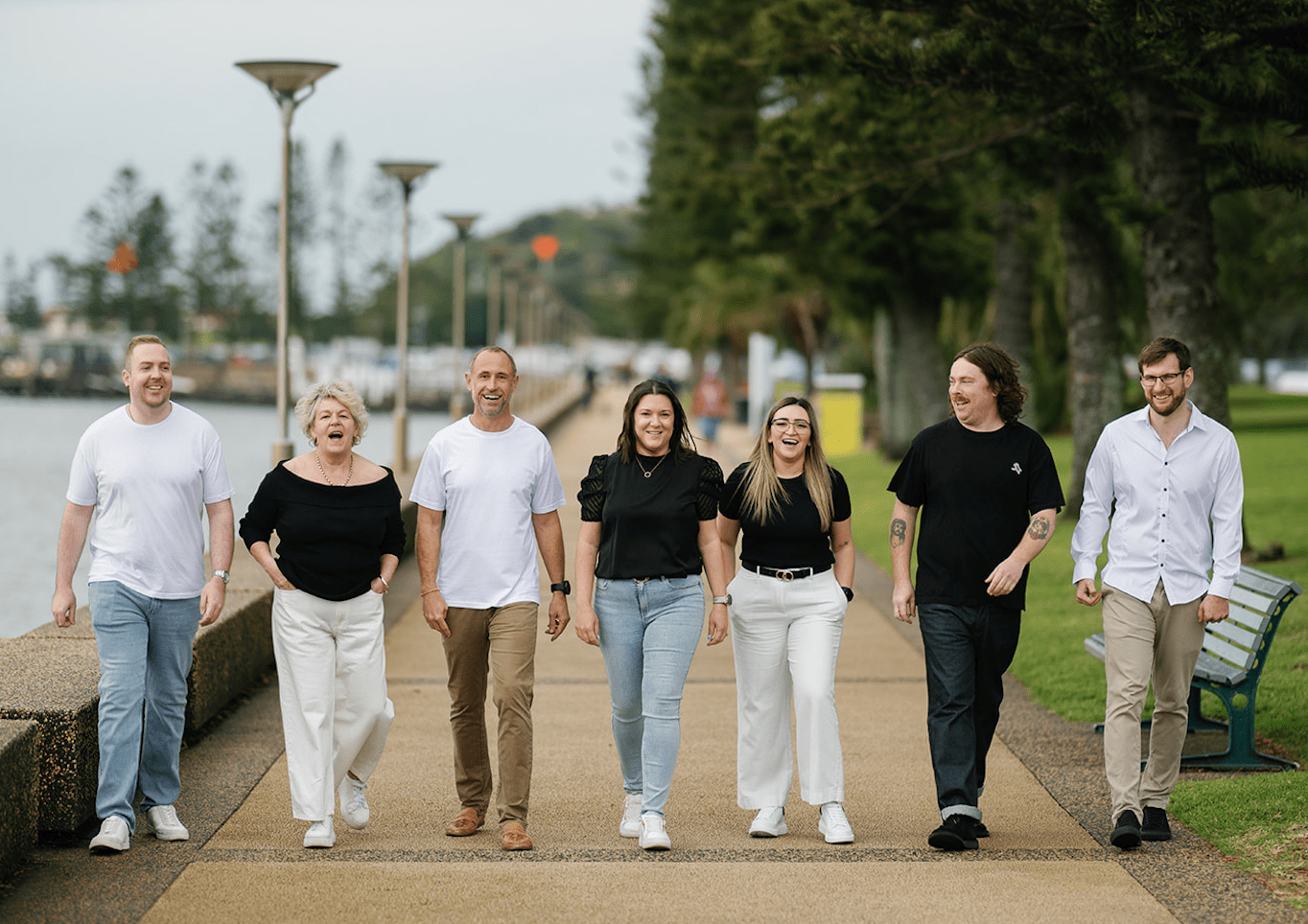 Seven people walk arm-in-arm on a path near water.  They are smiling and wearing casual clothes. Trees are in the background.