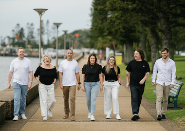 Group of seven people walking on a pathway near water; trees and a bench are visible.