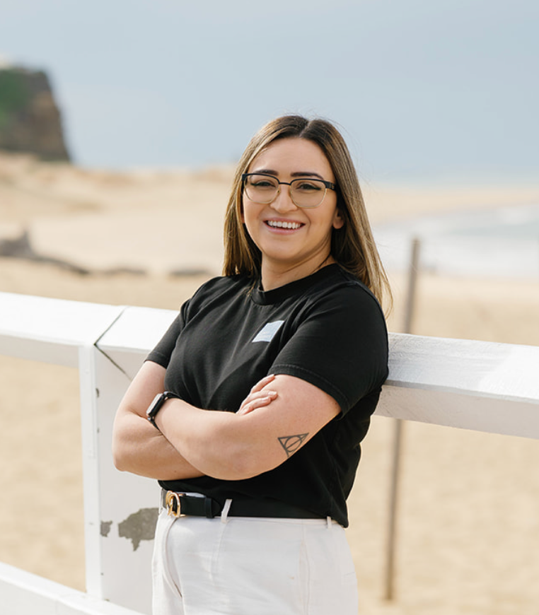 Woman with glasses, crossed arms, stands by a white fence on a beach, smiling.