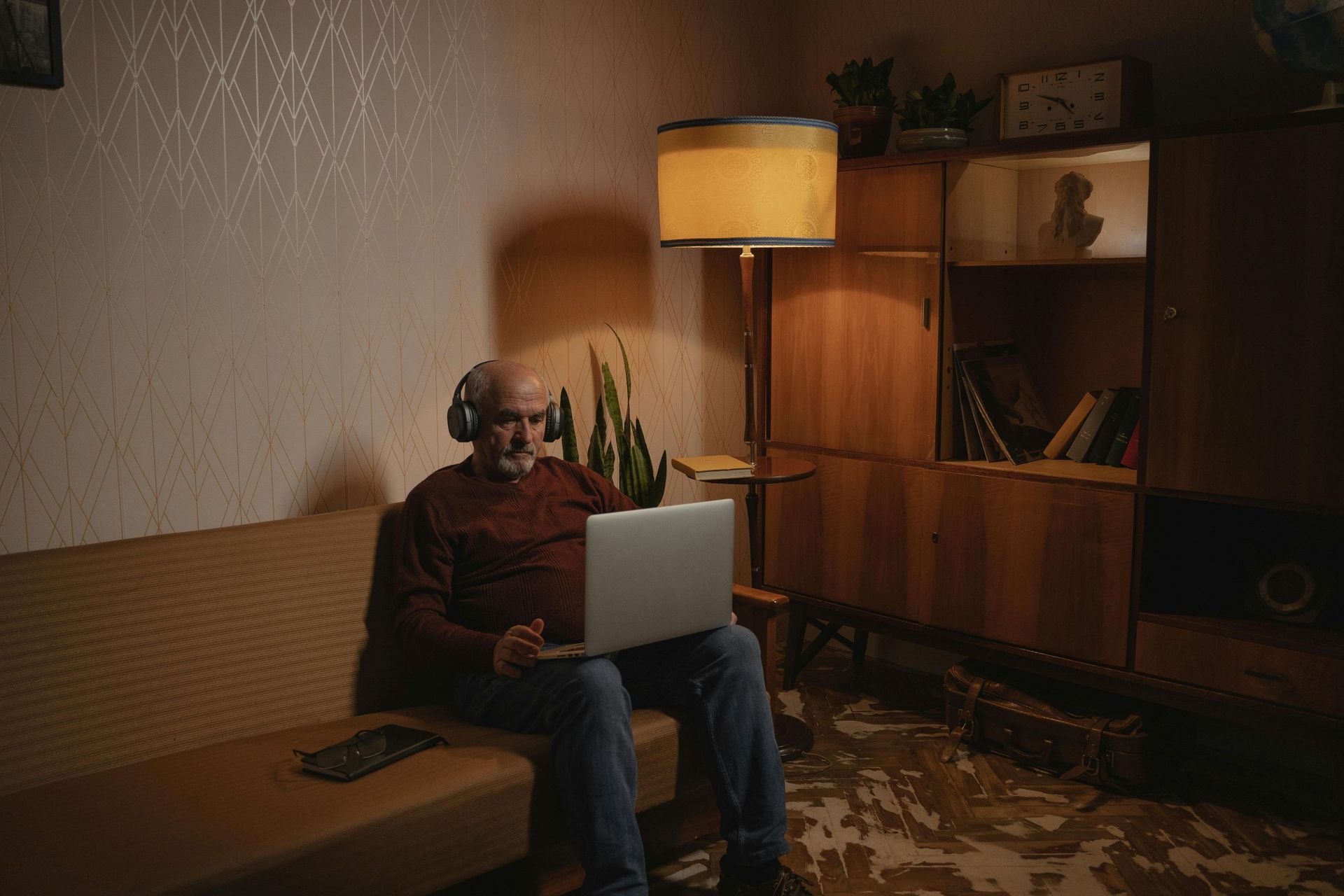 An elderly man sitting on a couch while working on a laptop and wearing headphones.