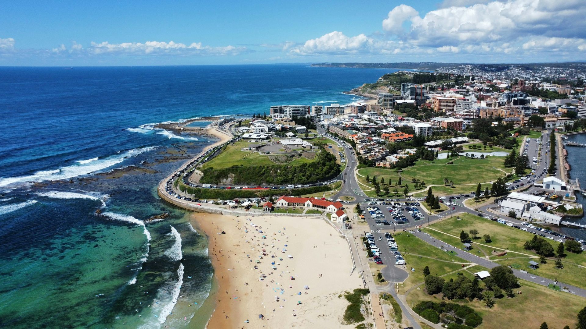 Aerial view of a coastal beach with turquoise water, green parkland, and city buildings along the shore.