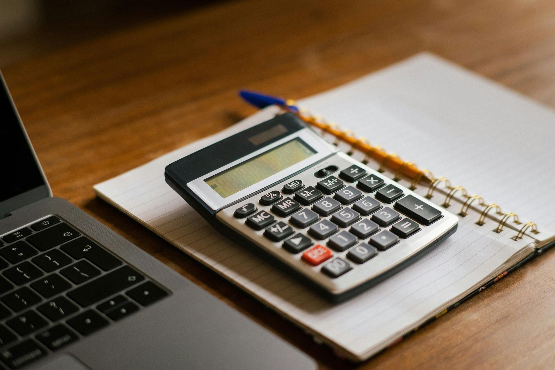 Calculator on a notebook beside a pencil and laptop on a wooden desk.