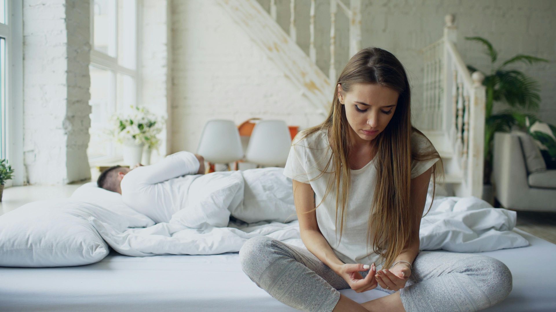 Woman sitting on a bed, looking down at a phone in a bright bedroom