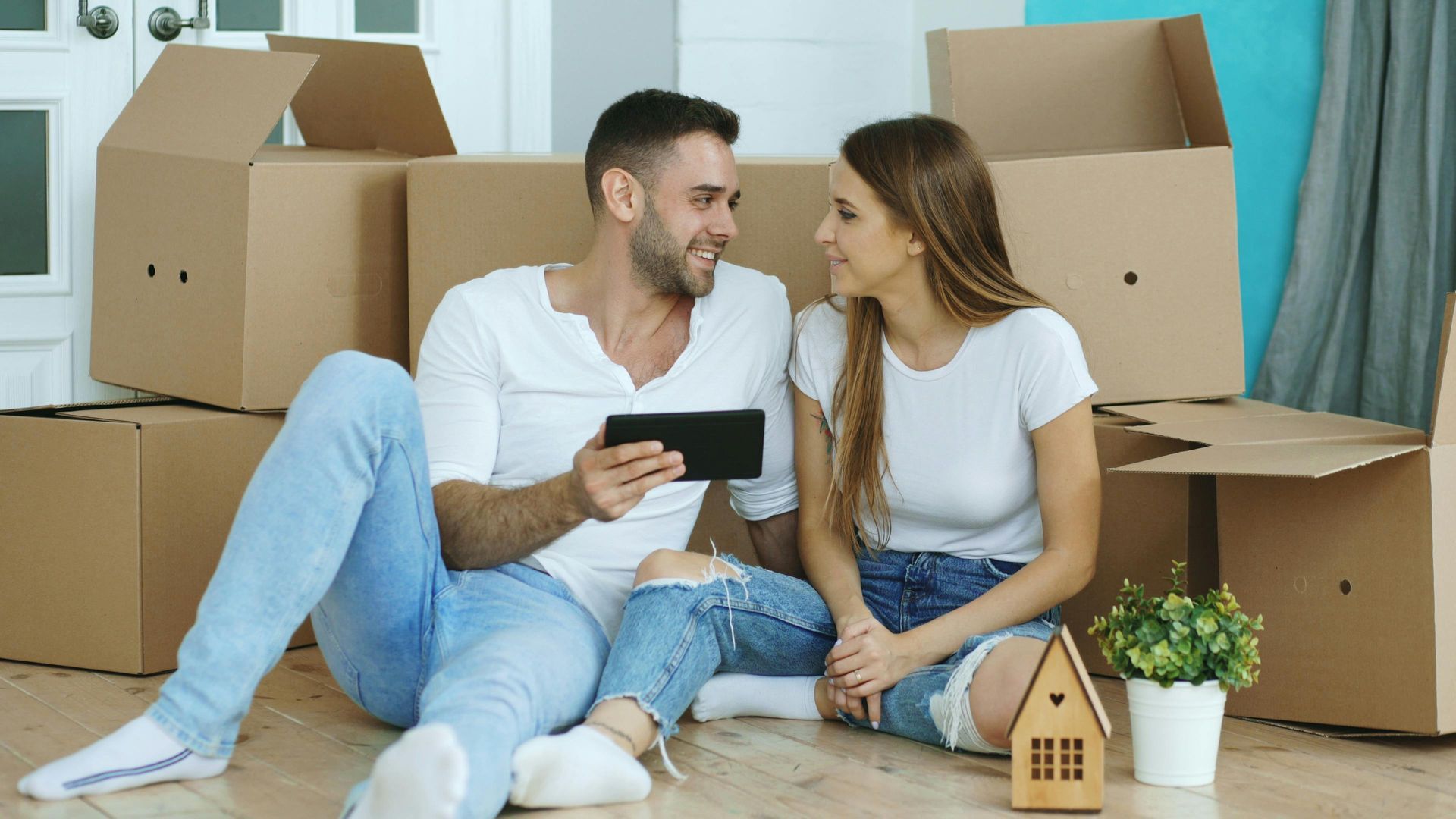 Couple sitting on the floor among moving boxes, looking at a phone in a new home.