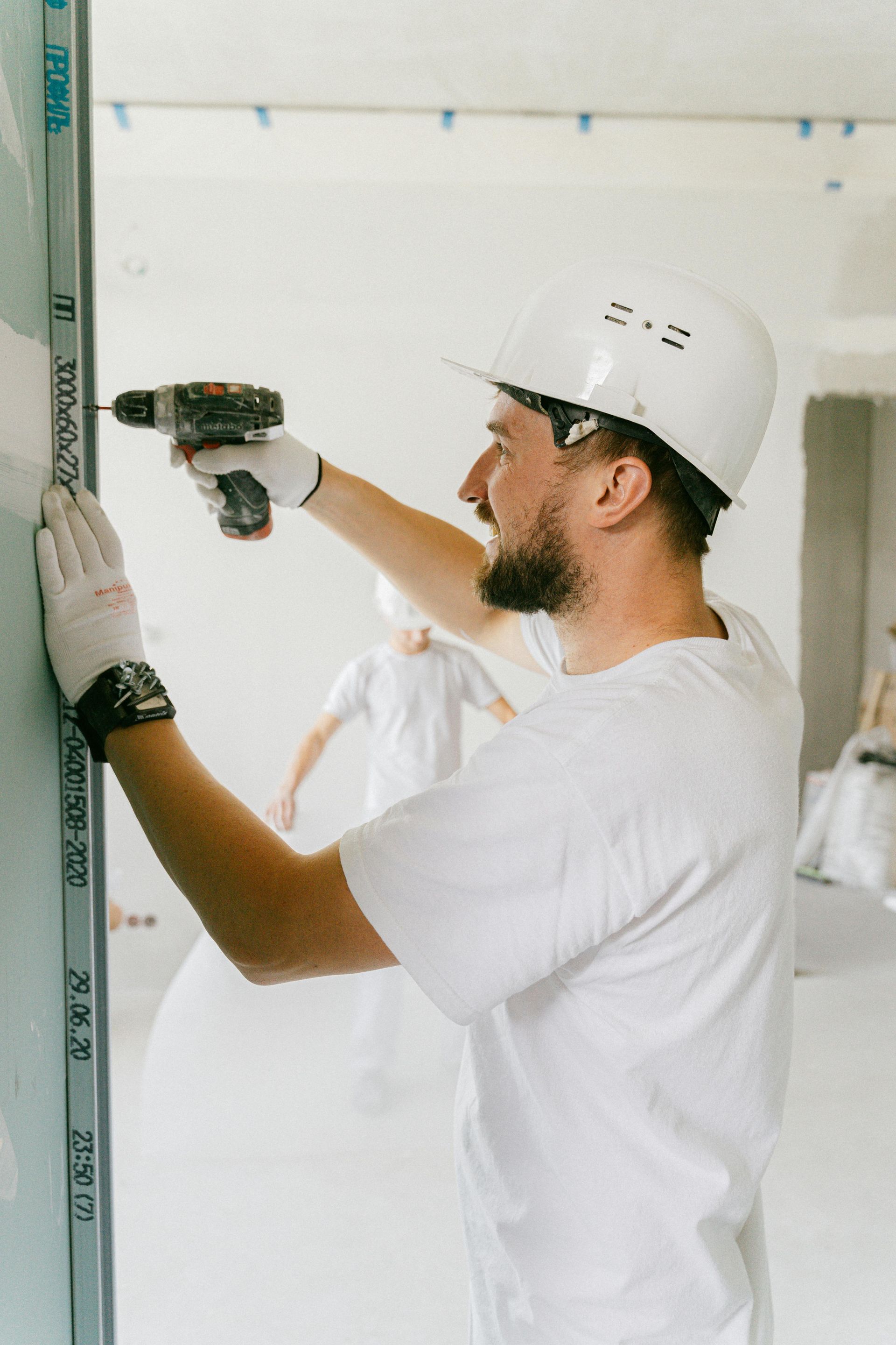 Worker in white hard hat using a power drill on a wall indoors