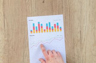 A hand points to a line graph on a paper with colorful bar graphs on a wooden table.