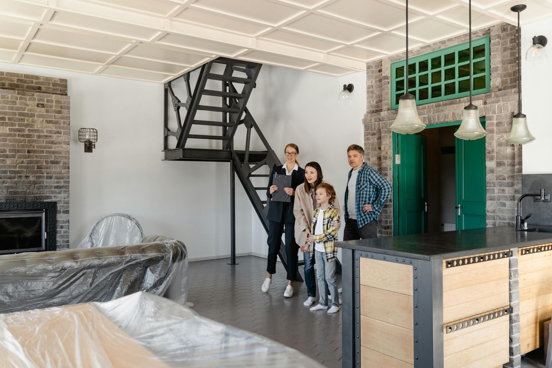 Four people standing in a modern room with a staircase, brick fireplace, and green pendant lights
