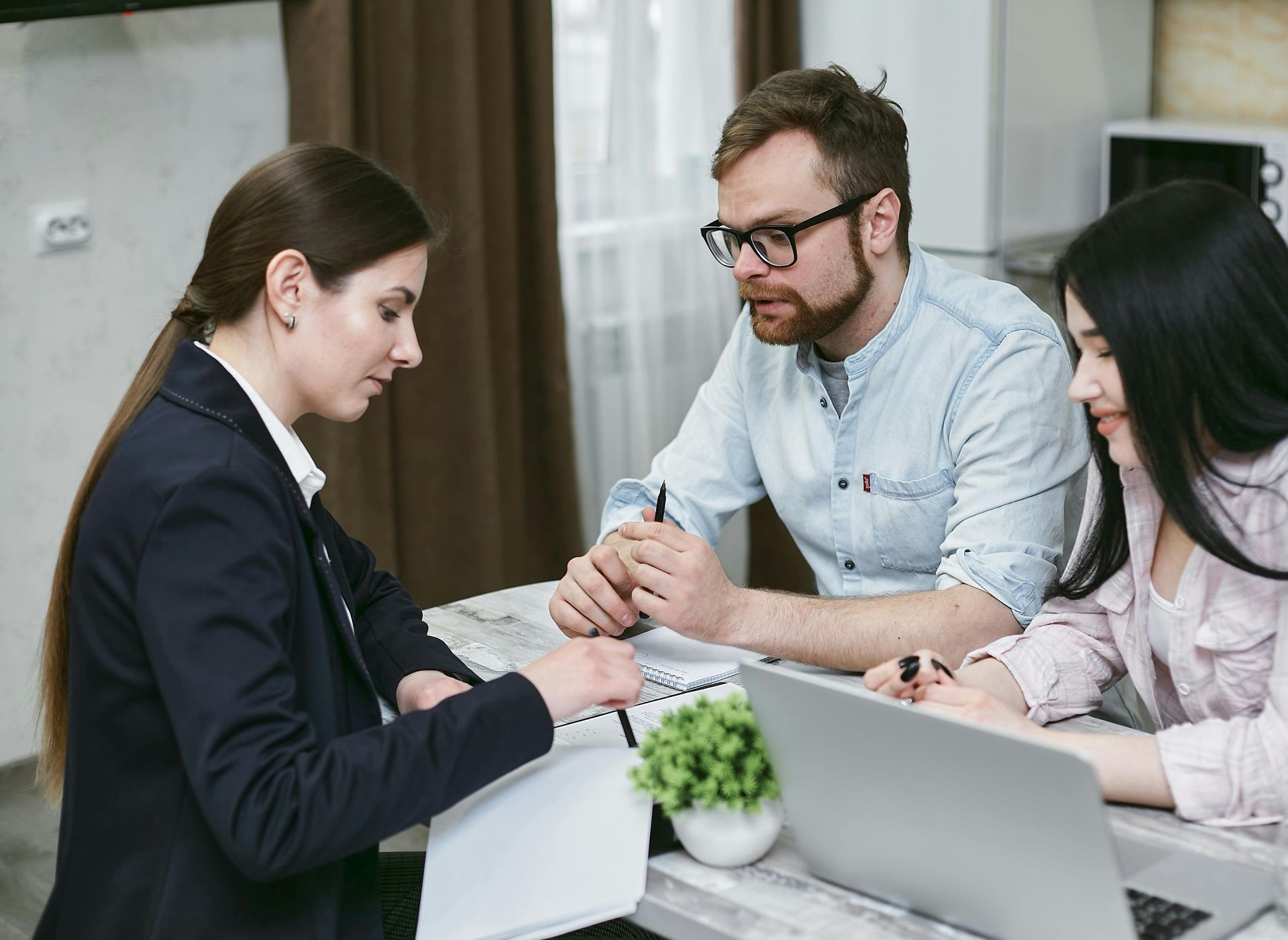 Three coworkers in a meeting, discussing notes around a laptop at a desk.