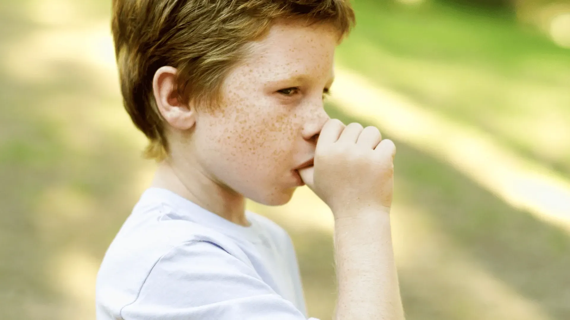 A young boy with red hair and freckles is biting his nails.