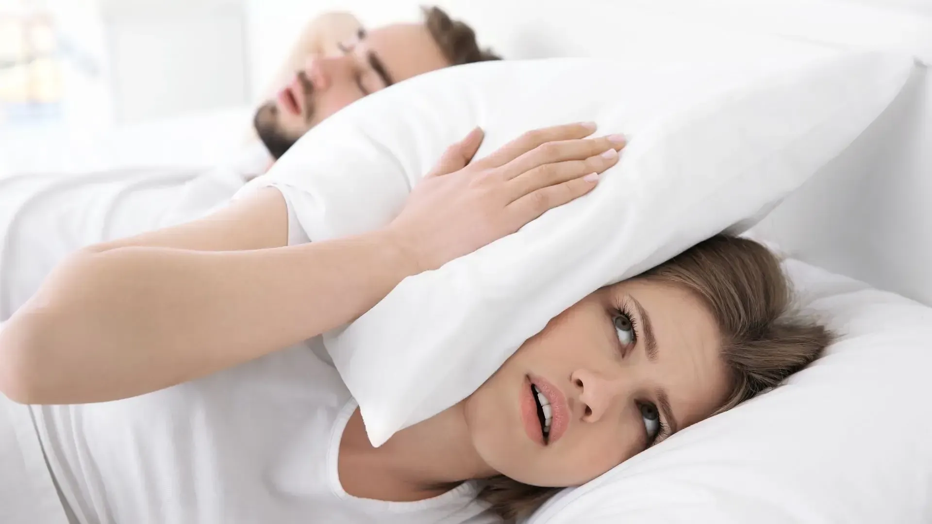 A woman is covering her ears with a pillow while a man sleeps next to her.