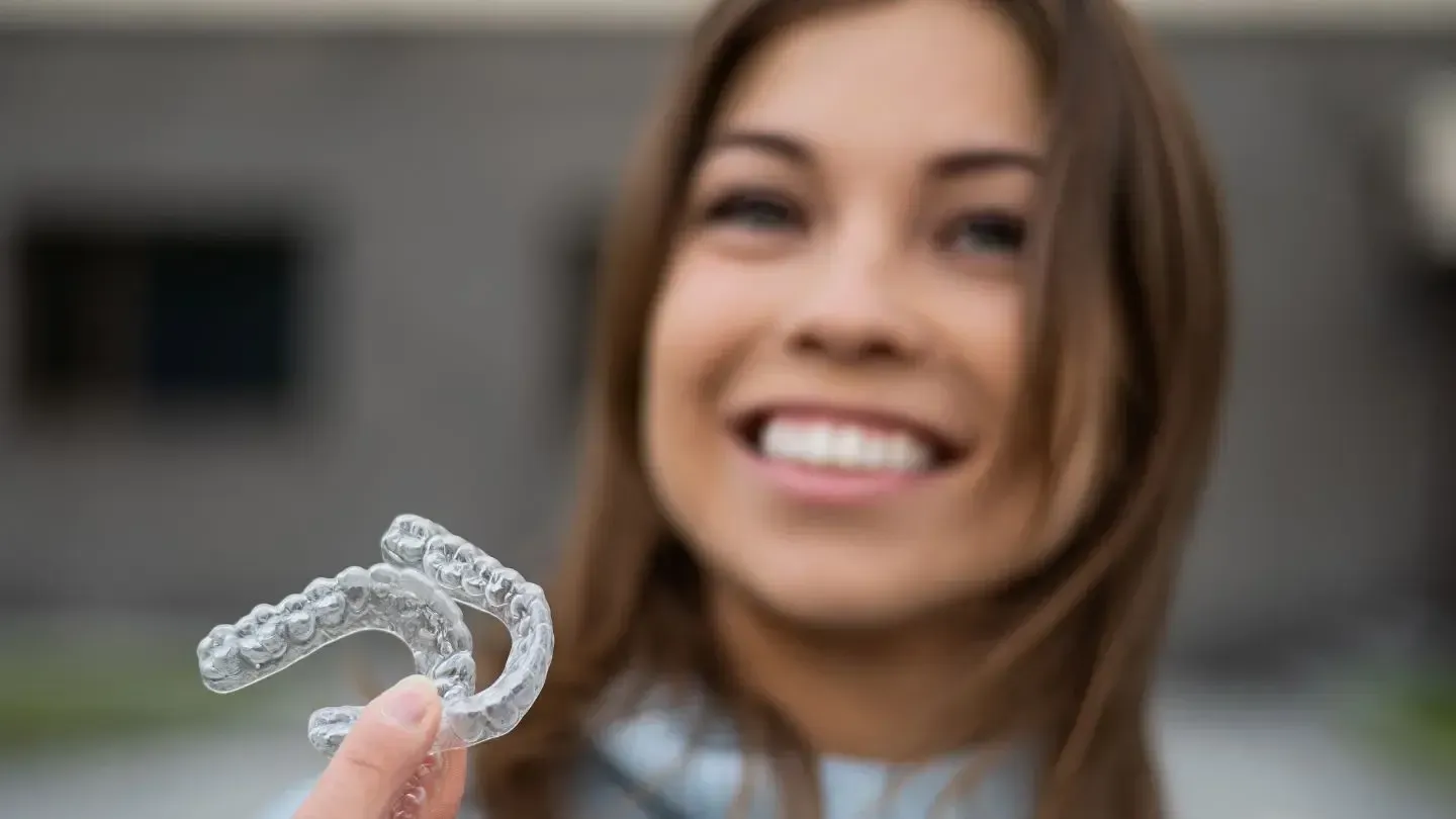 A woman is smiling while holding a clear mouth guard.
