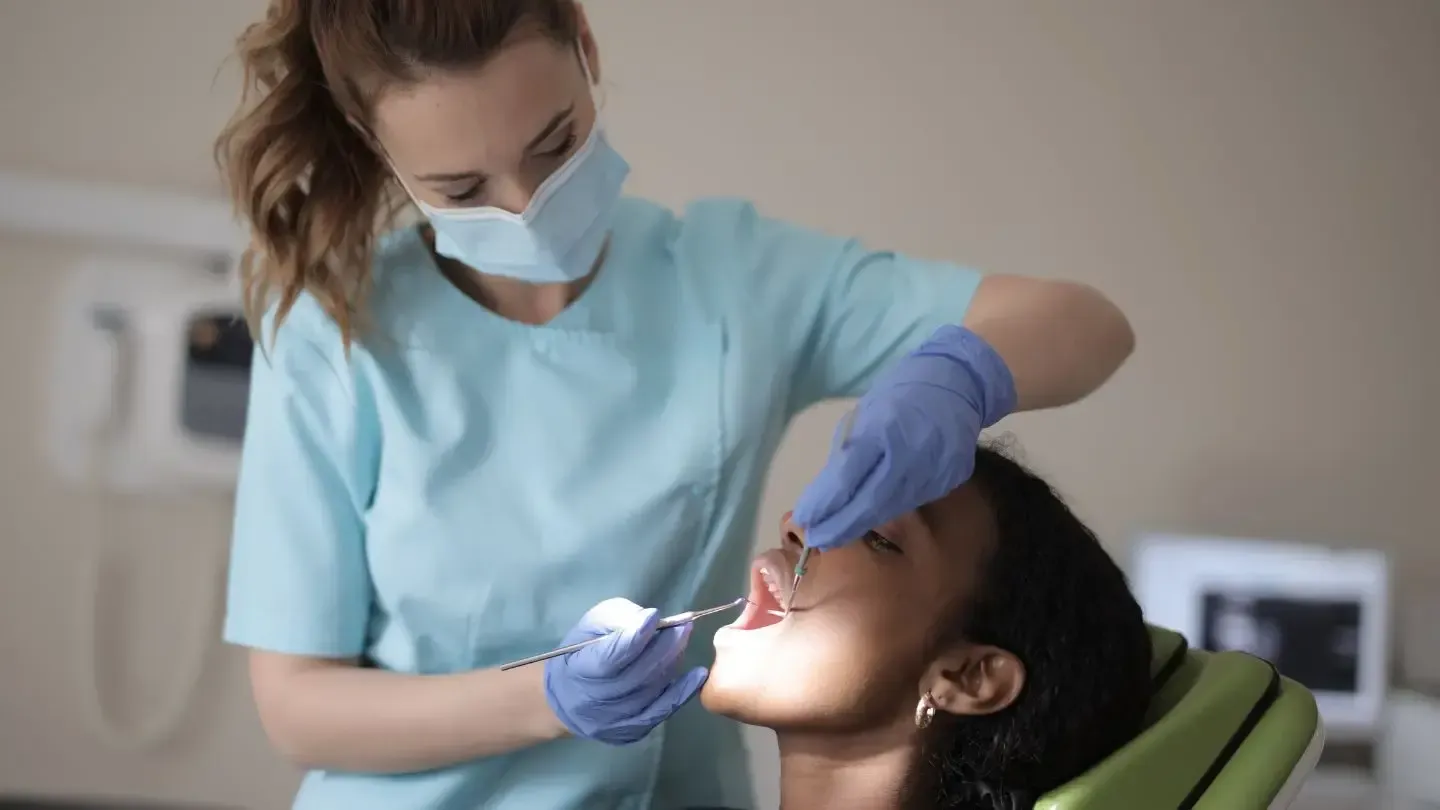 Dentist in mask and gloves examining a patient's teeth. Patient is sitting in a dental chair with mouth open.