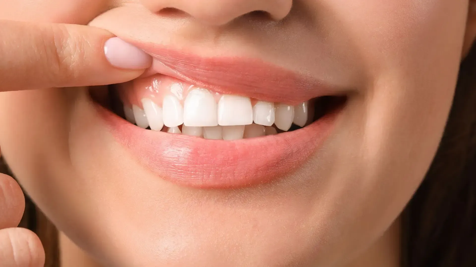 Close-up of a smiling woman with white teeth, pink lips, and a finger gently touching her upper lip.