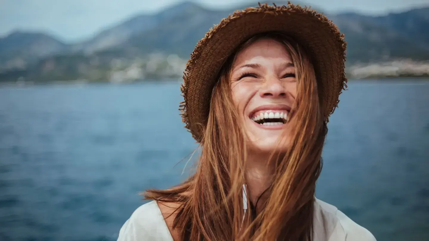 Woman with long brown hair wearing a straw hat laughs with ocean and mountains in the background.