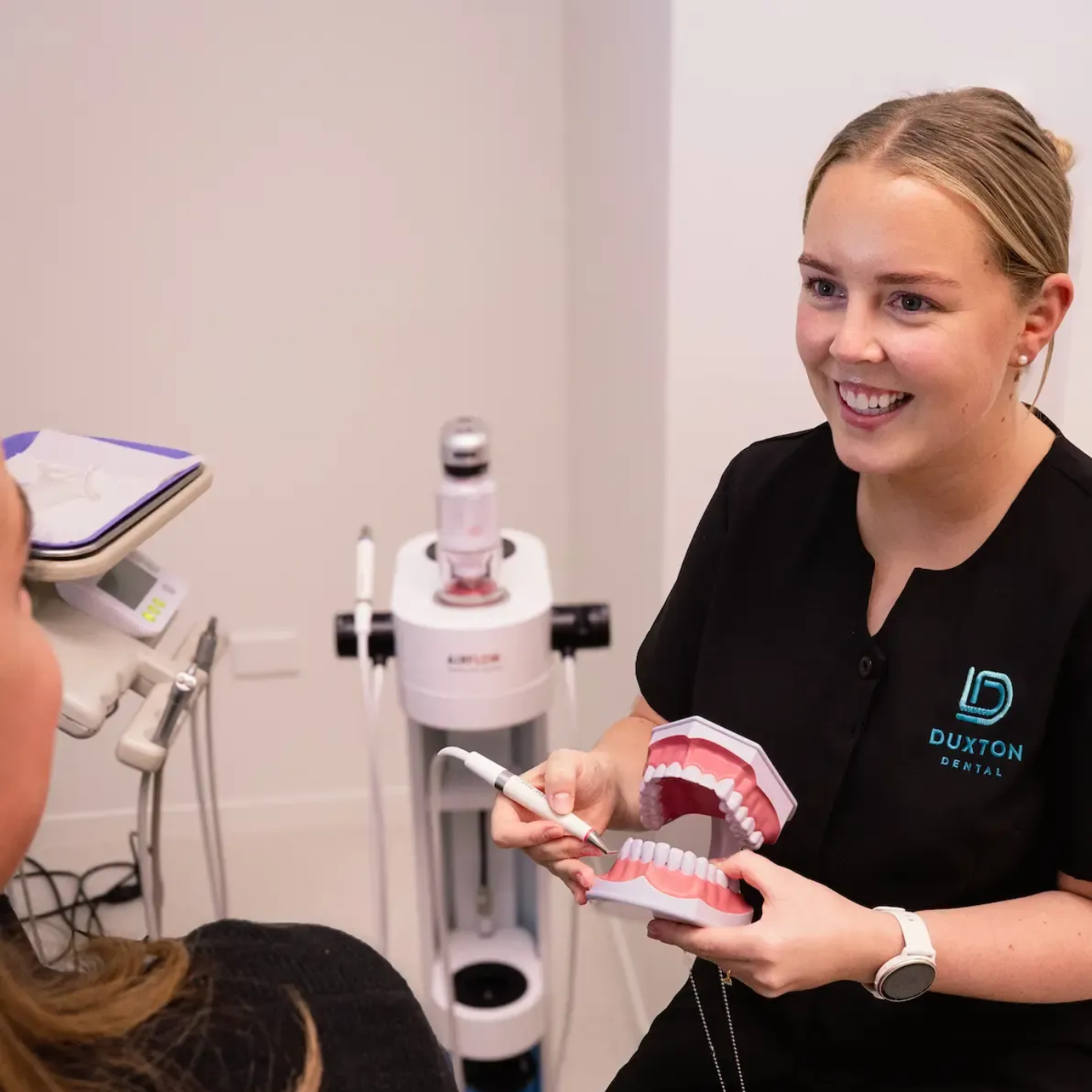 Dentist in black scrubs smiling, explaining with a model of teeth to a patient in a dental office.