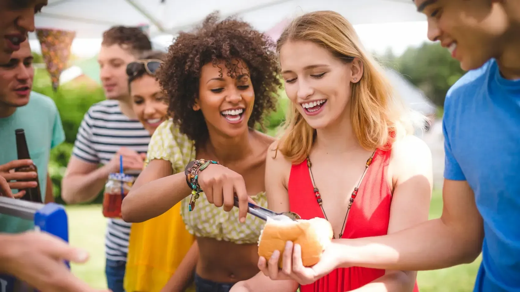 People at a gathering, two women smiling and scooping food onto a roll, others in the background.